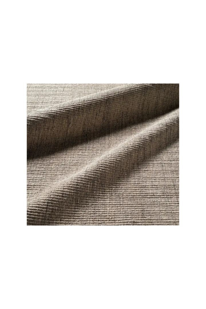 A close-up of textured woven area rug in a warm taupe-beige color, shown with soft folds and ribbed texture on a white background.