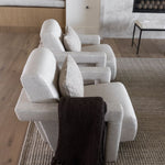 A row of modern light beige upholstered armchairs arranged on a woven Amity cream and gray area rug is ash wood in a contemporary living room, with a dark brown throw blanket draped over the front chair.