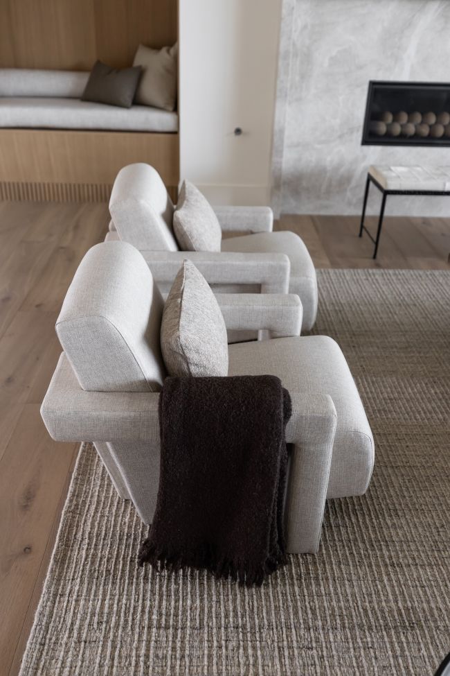 A row of modern light beige upholstered armchairs arranged on a woven Amity cream and gray area rug is ash wood in a contemporary living room, with a dark brown throw blanket draped over the front chair.