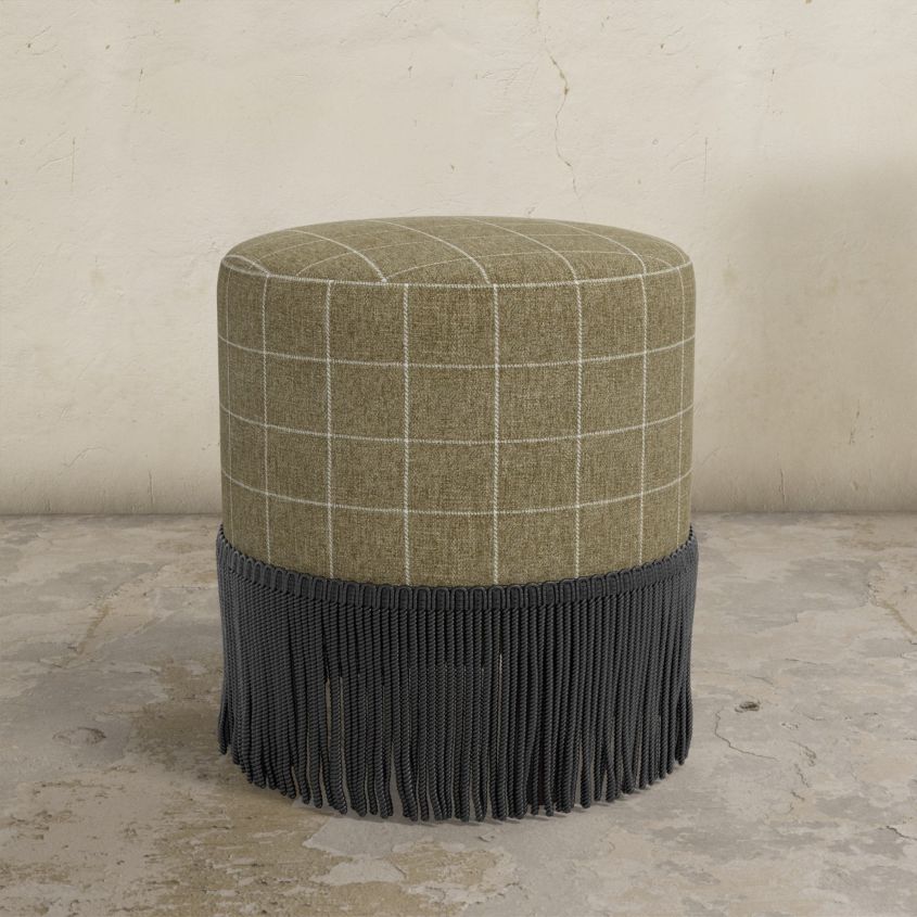 Round ottoman with plaid top and fringed bottom on a concrete floor.