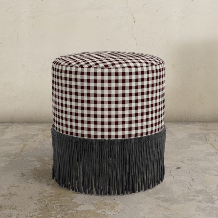 Gingham upholstered ottoman with fringe on a concrete floor.