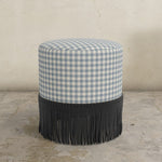 Round upholstered ottoman with blue and white gingham top and black fringed bottom on a concrete floor.