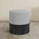 Round upholstered ottoman with blue and white gingham top and black fringed bottom on a concrete floor.