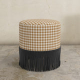 Golden gingham upholstered ottoman with black fringe on a concrete floor.