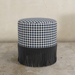 Round upholstered ottoman with gingham check and fringes on a concrete floor.