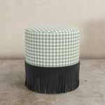 Round moss green gingham upholstered ottoman with black fringes on a concrete floor.
