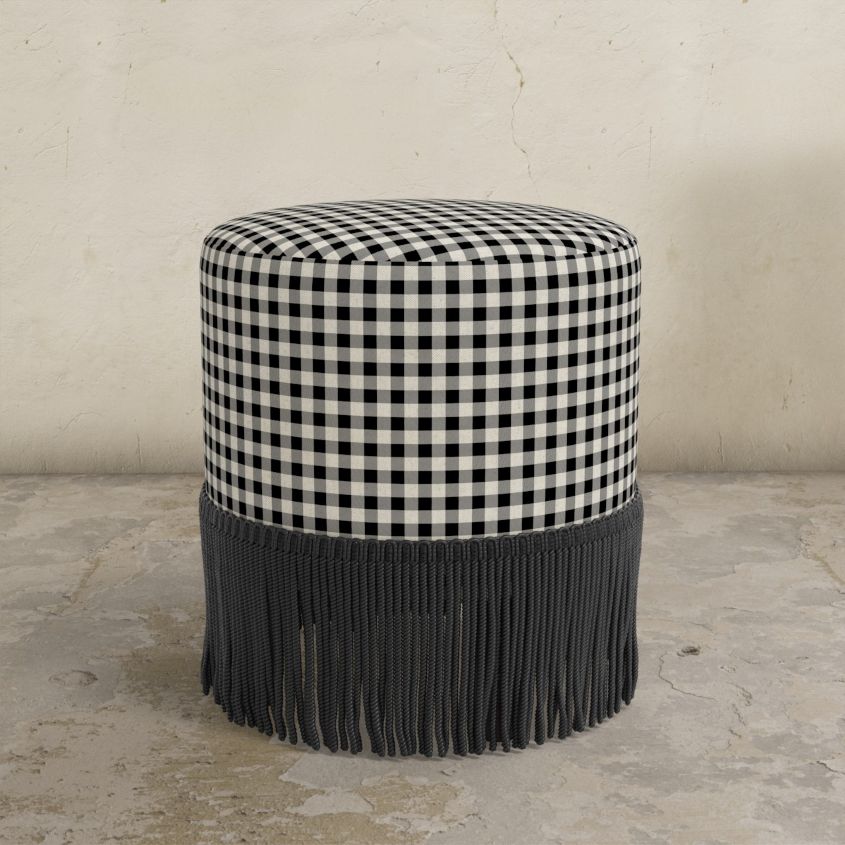 Black gingham round upholstered ottoman with fringed bottom on a concrete floor.