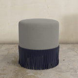 Gray upholstered ottoman with fringe on a concrete floor.