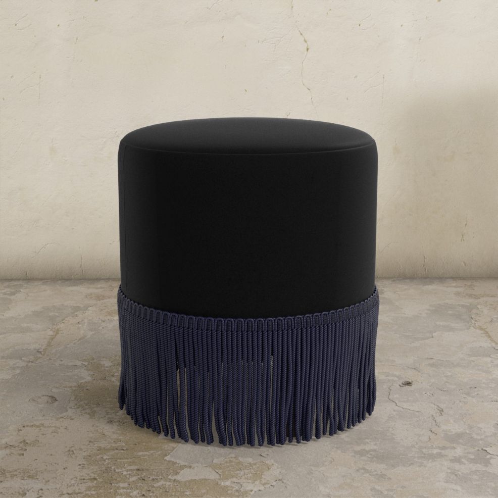 Black upholstered ottoman with fringes on a textured floor and light gray wall background