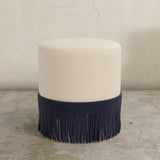 Round upholstered ottoman with black fringe on a textured floor.