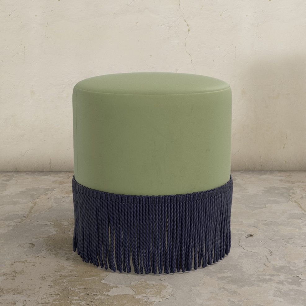 Green velvet upholstered ottoman with navy blue fringe on a stone floor.