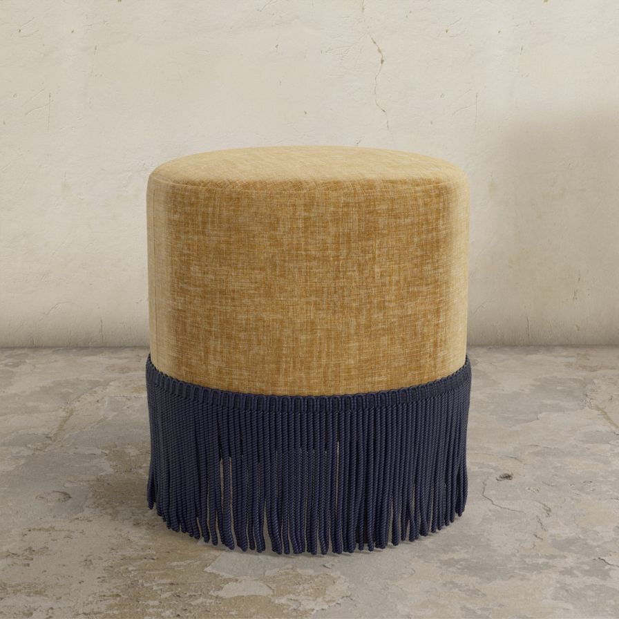 Round upholstered ottoman with beige top and navy fringes on a concrete floor.