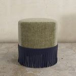 Round velvet upholstered ottoman with green top and navy fringes on a concrete floor.