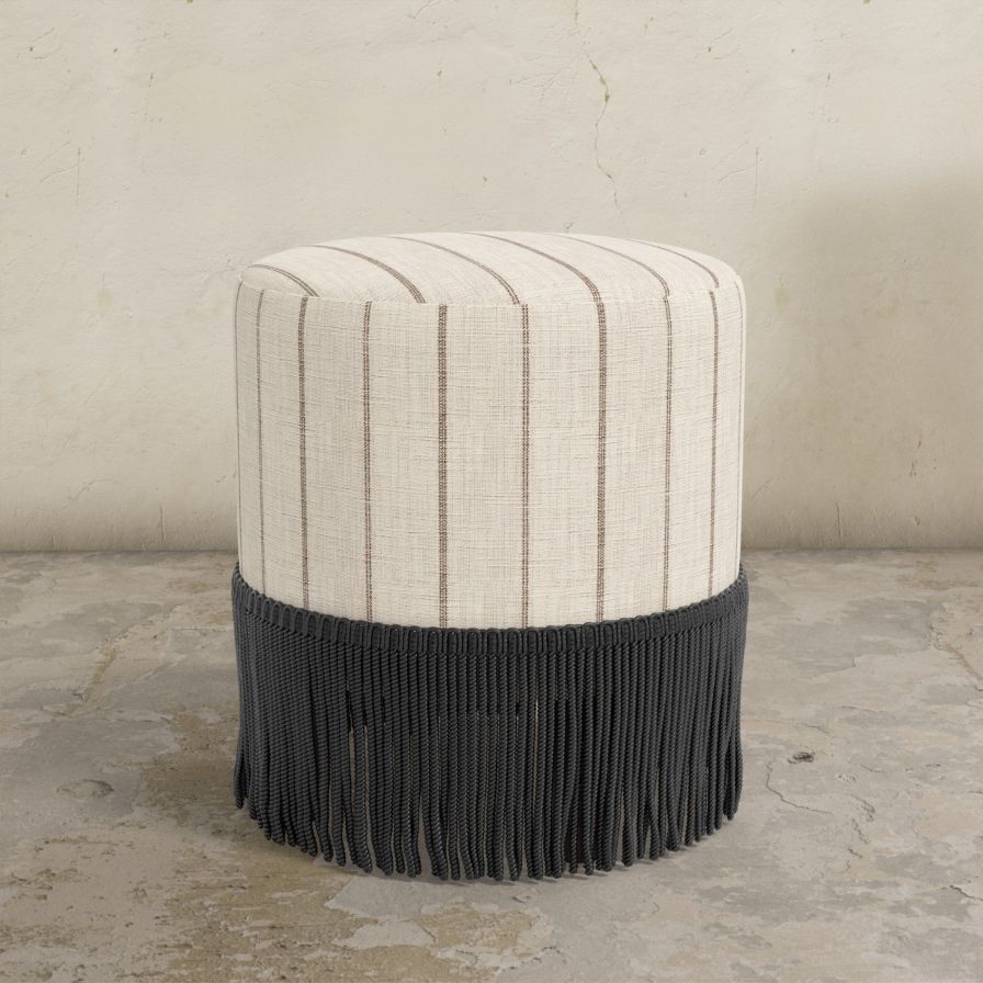 Round upholstered ottoman with striped pattern in neutral tones and fringe on a concrete floor.