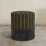 Brown upholstered ottoman with fringe on a concrete floor and light gray wall background