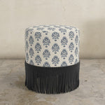 Round upholstered ottoman with pattern and black fringe on a concrete floor.
