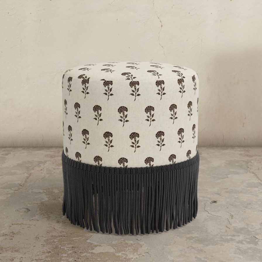 Round upholstered ottoman with pattern and black fringe on a textured floor.