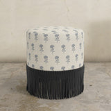 Round upholstered ottoman with blue flower and black fringed bottom on a concrete floor