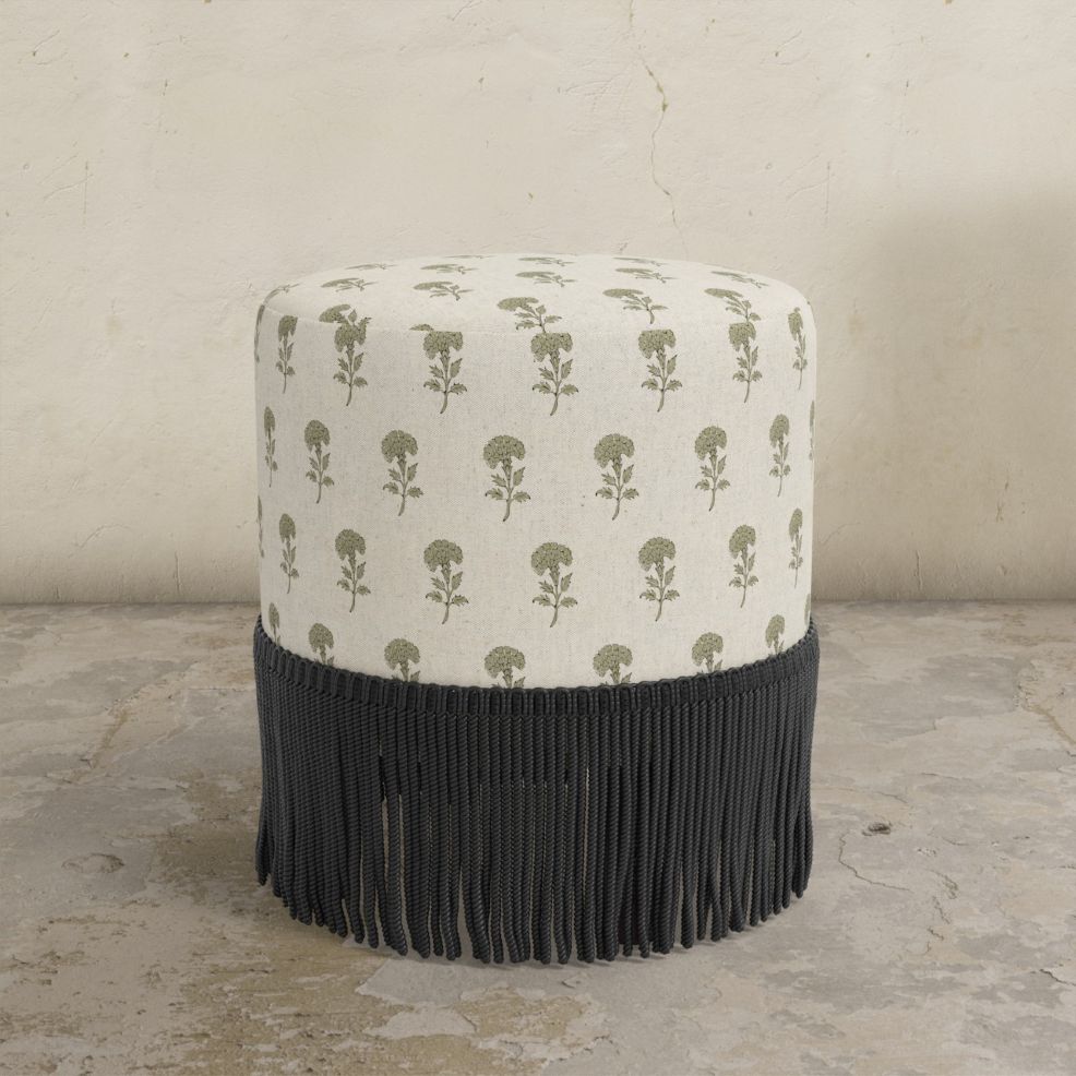 Round upholstered ottoman with patterened top and black fringes on a concrete floor.