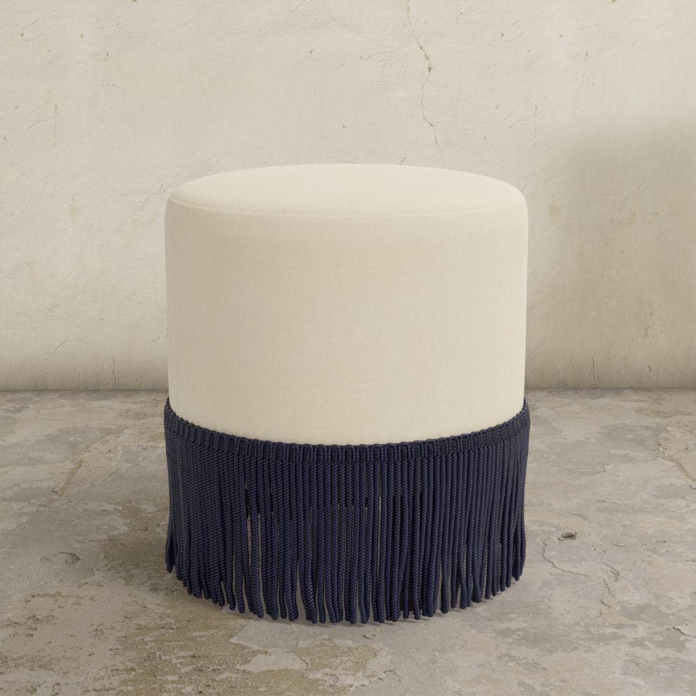 Round natural upholstered ottoman with fringe on a concrete floor.