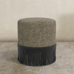 Round ottoman with textured upholstered top and black fringe bottom on a concrete floor.