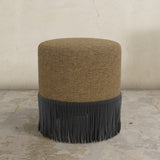 Round ottoman with brown top and black fringes on a stone floor.