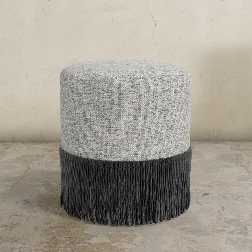 Round ottoman with textured gray top and black fringed bottom on a concrete floor.