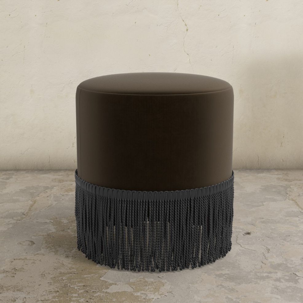 Round dark brown velvet upholstered ottoman with fringe detail on a textured floor.