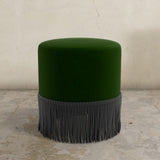 Green upholstered ottoman with gray fringe on a textured floor and light-colored wall background