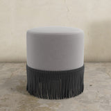 Gray upholstered ottoman with black fringe on a stone floor.