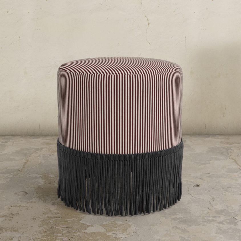 berry red striped upholstered  ottoman with fringe on a concrete floor.