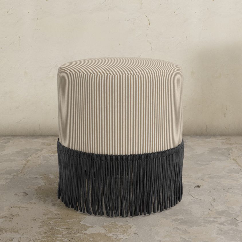 Striped upholstered ottoman with black fringe on a concrete floor.