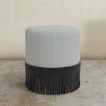 Round upholstered ottoman with blue and white striped top and fringed bottom on a concrete floor.