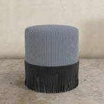 indigo blue striped upholstered ottoman with fringed bottom on a concrete floor