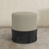 Round grey striped upholstered ottoman with black fringes on a concrete floor.