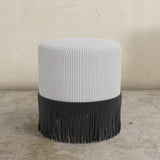 Striped upholstered ottoman with fringe on a concrete floor.