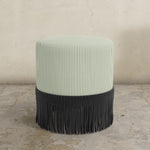 Moss green striped upholstered ottoman with fringe on a concrete floor.