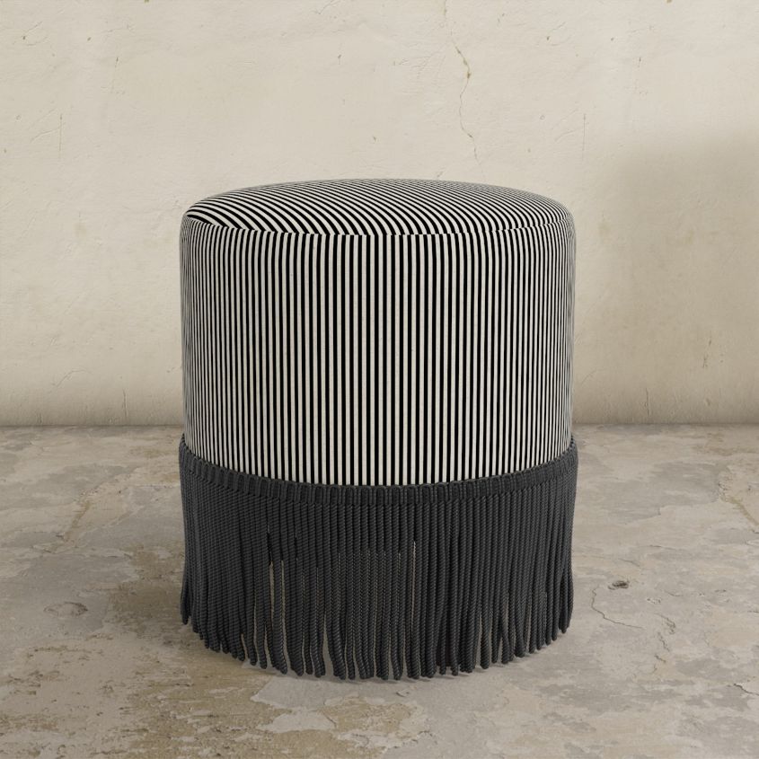 Black striped upholstered ottoman with fringes on a concrete floor against a textured wall.