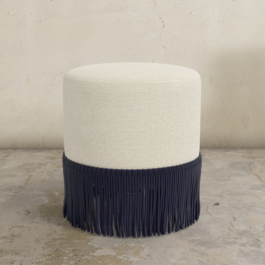 Round upholstered ottoman with white top and navy fringes on a concrete floor.