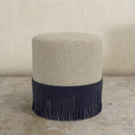 Round upholstered ottoman with beige top and black fringes on a concrete floor.