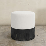 Round upholstered ottoman with white cushion and fringes on a concrete floor.