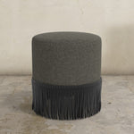 Gray upholstered ottoman with fringe on a concrete floor.
