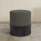 Gray upholstered ottoman with fringe on a concrete floor.
