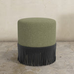 Green upholstered ottoman with black fringe on a concrete floor.