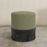 Green upholstered ottoman with black fringe on a concrete floor.