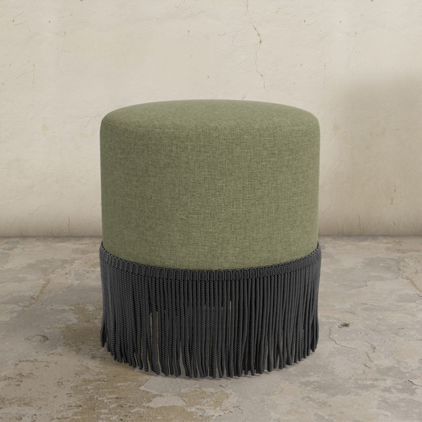 Green upholstered ottoman with black fringe on a concrete floor.