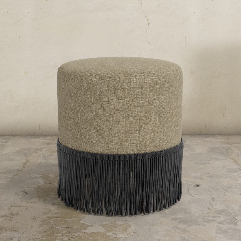 Round upholstered ottoman with beige top and black fringes on a concrete floor.