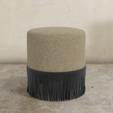 Round upholstered ottoman with beige top and black fringes on a concrete floor.