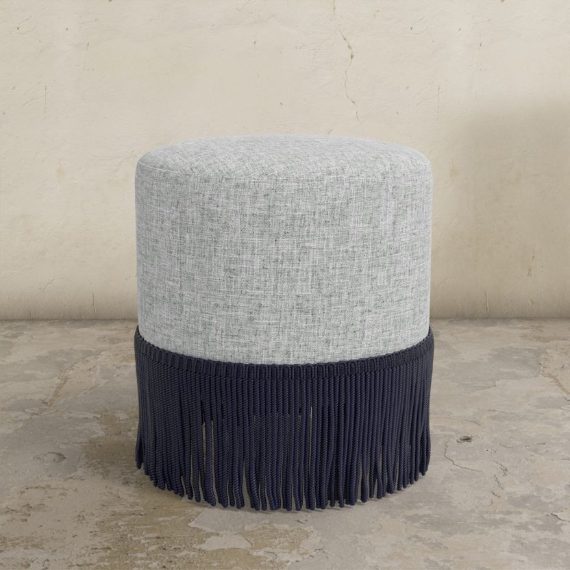 Round upholstered ottoman with gray top and navy fringes on a concrete floor.