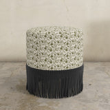 Round upholstered ottoman with floral pattern and black fringe on a concrete floor.